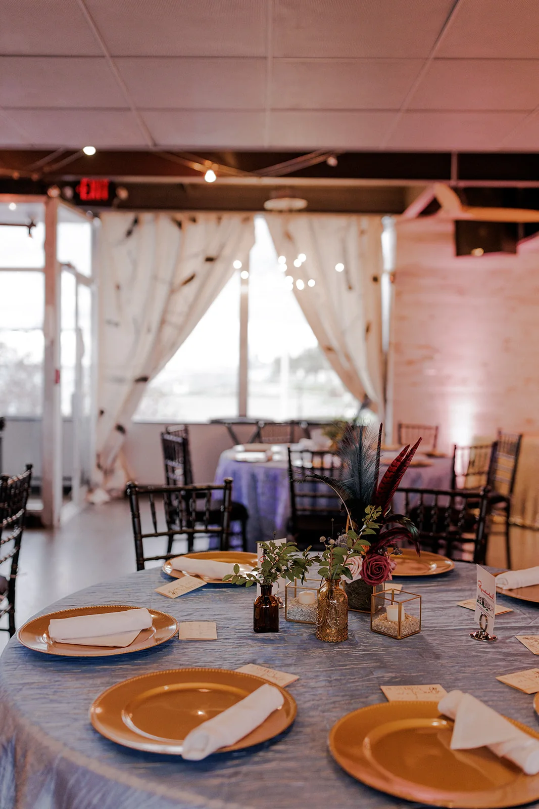 The Club set for a wedding reception with round tables, gold chargers and black Chiavari chairs, the water in the windows beyond