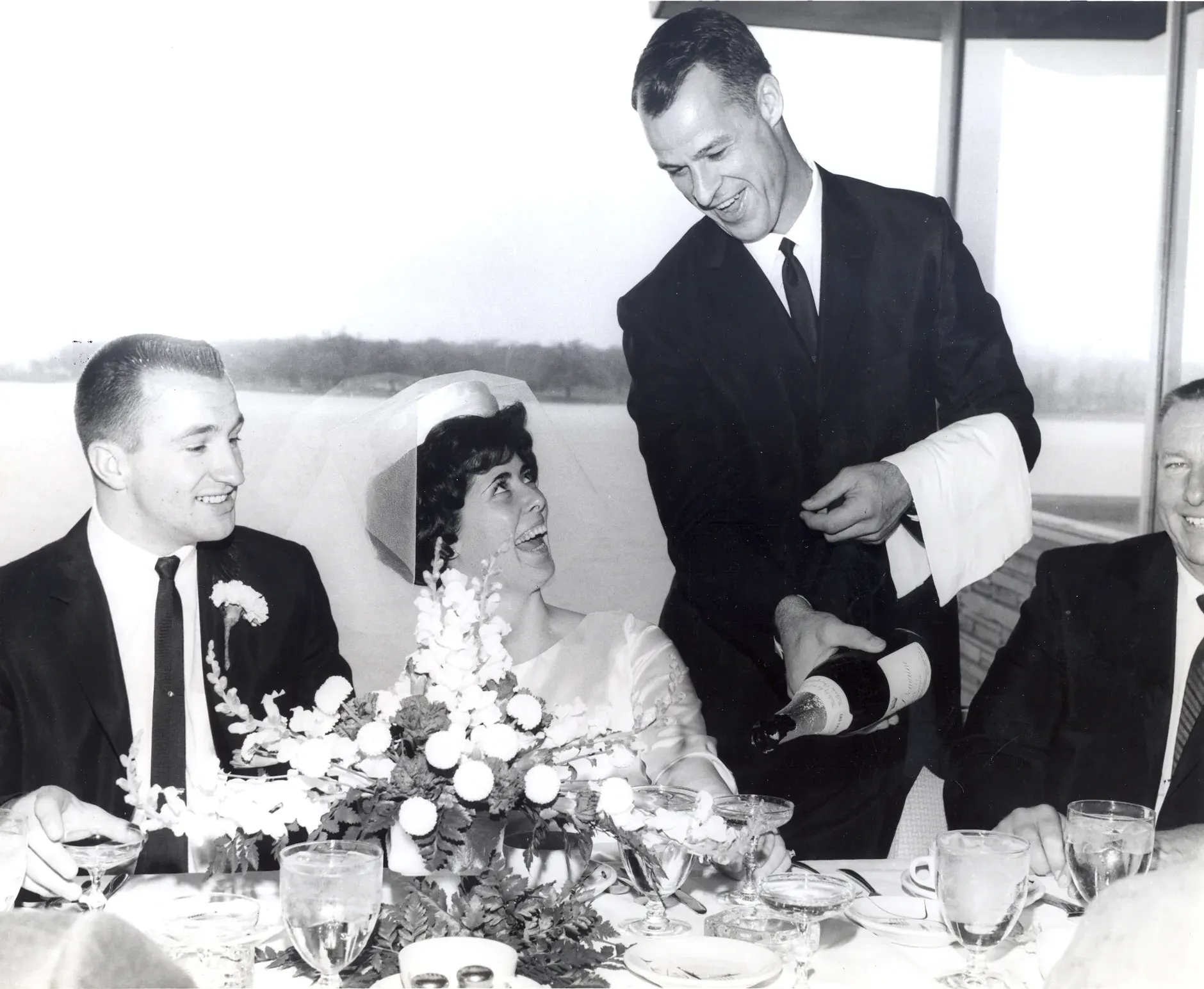 Gordie Howe in a dark suit pours champagne for a bride in a veil and her groom at their table, river light in the windows behind them.