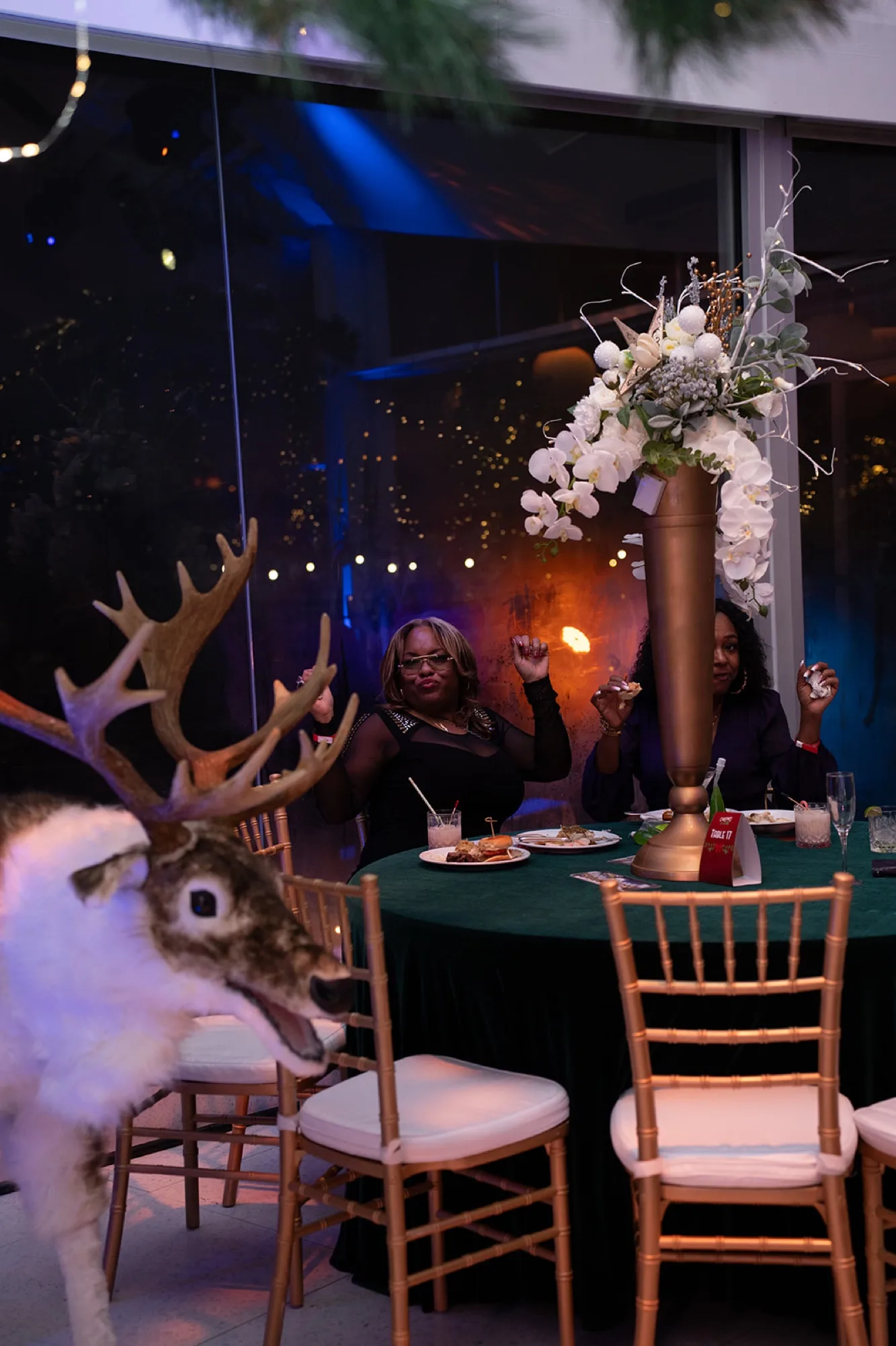A round table set with green linens, a white floral centerpiece and a reindeer figure by the window